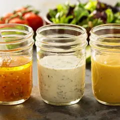 Trzy słoiki z domowymi sosami do sałatek: vinaigrette, ziołowy i musztardowy. Idealne, by dowiedzieć się, jak zrobić sos do sałatki z oleju.