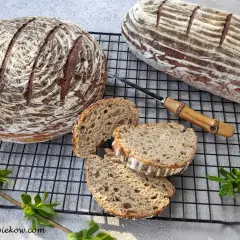 Dwa bochenki chleba żytniego na zakwasie, jeden okrągły, drugi podłużny, z kilkoma kromkami obok.