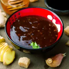 Sos teriyaki przepis: miseczka pełna aromatycznego sosu, obok świeży imbir, czosnek i brązowy cukier. Idealne składniki do domowego sosu teriyaki.