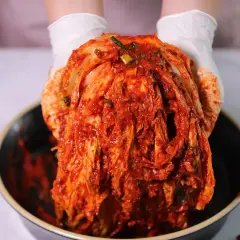 Ręce w białych rękawiczkach trzymają całą, czerwoną kapustę kimchi nad miską. Gdzie kupić kapustę kimchi?