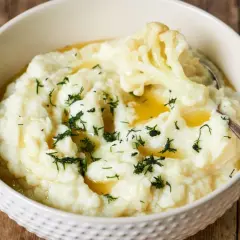 Aksamitne puree z kalafiora, polane masłem i posypane koperkiem, w białej misce na drewnianym tle.
