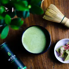 Miska z matchą, bambusowa miotełka, puszka MOYA Matcha i czekolada na drewnianym tle.