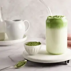 Zielony napój z mlekiem i sproszkowaną matchą, który idealnie oddaje, jak smakuje matcha latte. Obok miseczka z matchą i łyżeczka.
