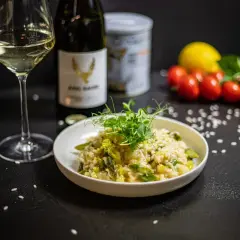 Klasyczne risotto z kurczakiem, podane z zielonymi szparagami i kiełkami, obok kieliszka białego wina i butelki.