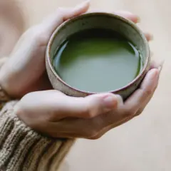 Dłonie w beżowym swetrze trzymają miseczkę z zielonym napojem. Czy matcha to zielona herbata? Tak, to sproszkowana zielona herbata.