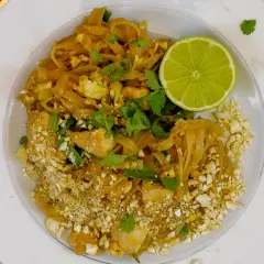 Talerz z Pad Thai, posypany orzeszkami ziemnymi, z połówką limonki i kolendrą.