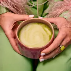 Dłonie trzymają kubek z zieloną matchą, zastanawiając się, czy matcha odchudza. Ubrana w pistacjowy strój z różowymi piórami.