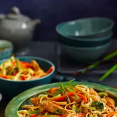 Makaron Chow Mein z kurczakiem i warzywami w misce, na tle czajniczka i pałeczek.