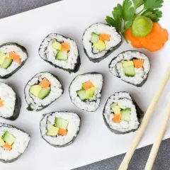 Talerz z rolkami sushi z awokado, ogórkiem i marchewką. Obok zielona pasta wasabi i ozdobna marchewka. Pałeczki czekają, by zacząć ucztę z czym sushi.