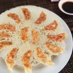Chrupiące pierożki gyoza ułożone na talerzu, z delikatną, złotą panierką z ciasta. Obok miseczka sosu sojowego i pałeczki.