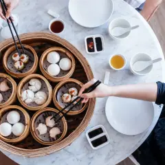 Różnorodne dim sum w bambusowych parownikach, gotowe do degustacji. Ręka z pałeczkami wybiera jeden z przysmaków.