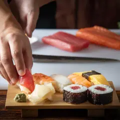 Ręce układają kawałek tuńczyka na sushi. Na desce widać też krewetkę, kalmara, łososia, omlet i rolki. Idealne składniki, by dodać do sushi.