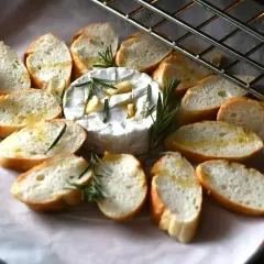 Pyszny, pieczony camembert z czosnkiem i rozmarynem, otoczony chrupiącymi grzankami skropionymi oliwą. Idealna przekąska.