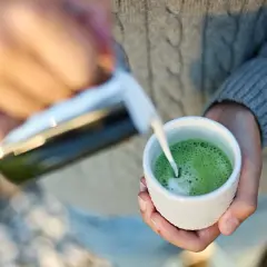 Ręce nalewają mleko do filiżanki z zieloną matchą.