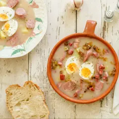 Dwa talerze z barszczem białym i żurkiem, pełne jajek, kiełbasy i ziemniaków. Obok kromka chleba i czosnek.