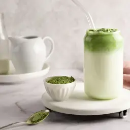 Zielony napój z mlekiem i sproszkowaną matchą, który idealnie oddaje, jak smakuje matcha latte. Obok miseczka z matchą i łyżeczka.