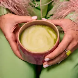 Dłonie trzymają kubek z zieloną matchą, zastanawiając się, czy matcha odchudza. Ubrana w pistacjowy strój z różowymi piórami.