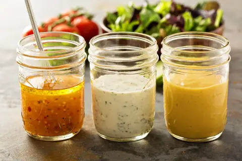 Trzy słoiki z domowymi sosami do sałatek: vinaigrette, ziołowy i musztardowy. Idealne, by dowiedzieć się, jak zrobić sos do sałatki z oleju.