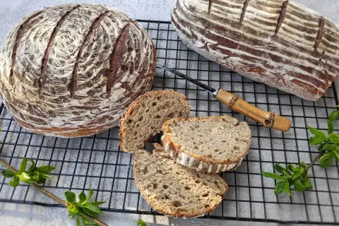 Dwa bochenki chleba żytniego na zakwasie, jeden okrągły, drugi podłużny, z kilkoma kromkami obok.