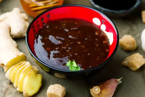 Sos teriyaki przepis: miseczka pełna aromatycznego sosu, obok świeży imbir, czosnek i brązowy cukier. Idealne składniki do domowego sosu teriyaki.
