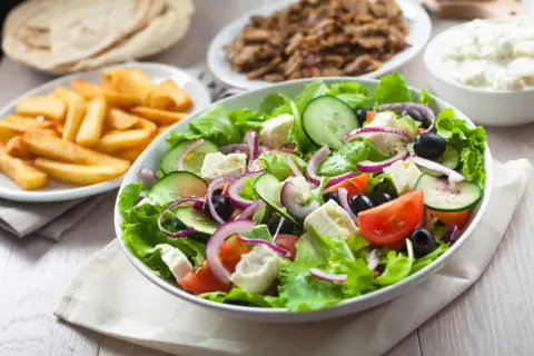 Świeża sałatka z sałatą, pomidorami, ogórkiem, fetą i oliwkami – idealne co do sałatki gyros. Obok frytki i mięso gyros.