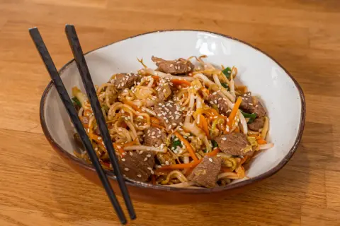 Makaron Chow Mein z wołowiną, warzywami i sezamem w misce z pałeczkami.