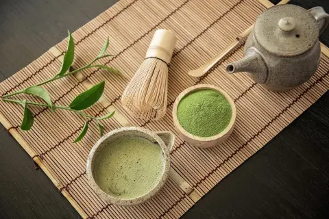 Tradycyjne akcesoria do parzenia matcha: bambusowa miotełka, czarka z napojem, drewniana miseczka z zielonym proszkiem.