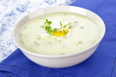 Kremowa zupa z sałaty z jajkiem i ziołami, podana w białej misce na niebieskim obrusie.