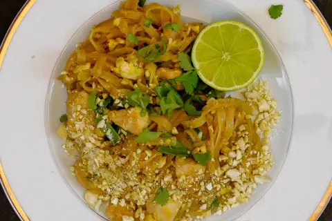 Talerz z Pad Thai, posypany orzeszkami ziemnymi, z połówką limonki i kolendrą.