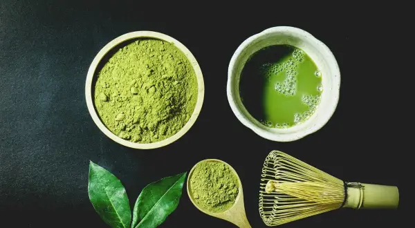 Miska z zielonym proszkiem matcha, filiżanka napoju, drewniana łyżka i bambusowa miotełka.
