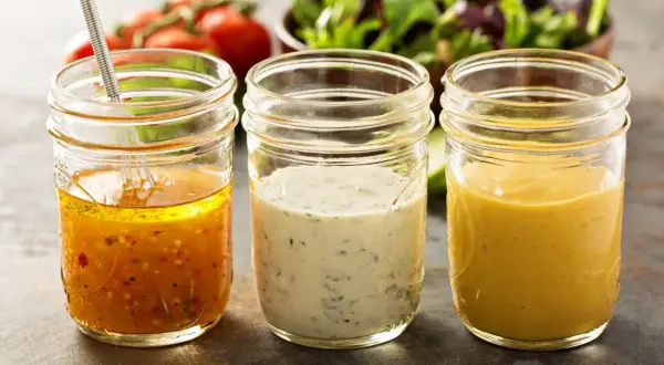 Trzy słoiki z domowymi sosami do sałatek: vinaigrette, ziołowy i musztardowy. Idealne, by dowiedzieć się, jak zrobić sos do sałatki z oleju.
