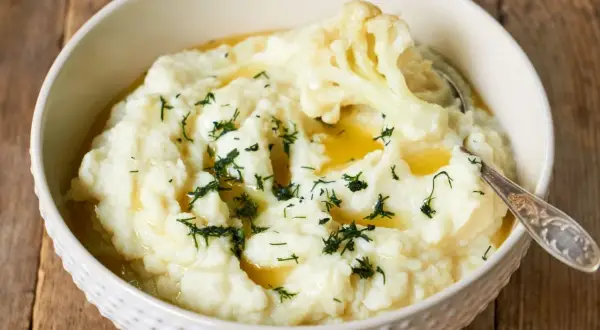 Aksamitne puree z kalafiora, polane masłem i posypane koperkiem, w białej misce na drewnianym tle.