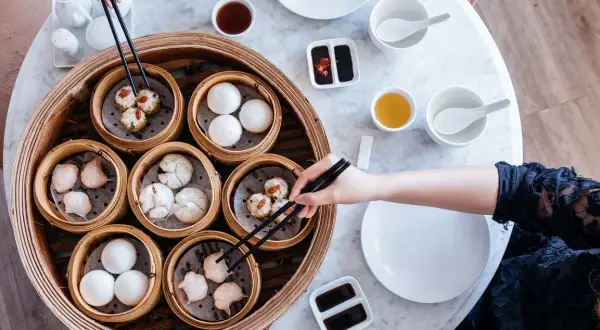 Różnorodne dim sum w bambusowych parownikach, gotowe do degustacji. Ręka z pałeczkami wybiera jeden z przysmaków.