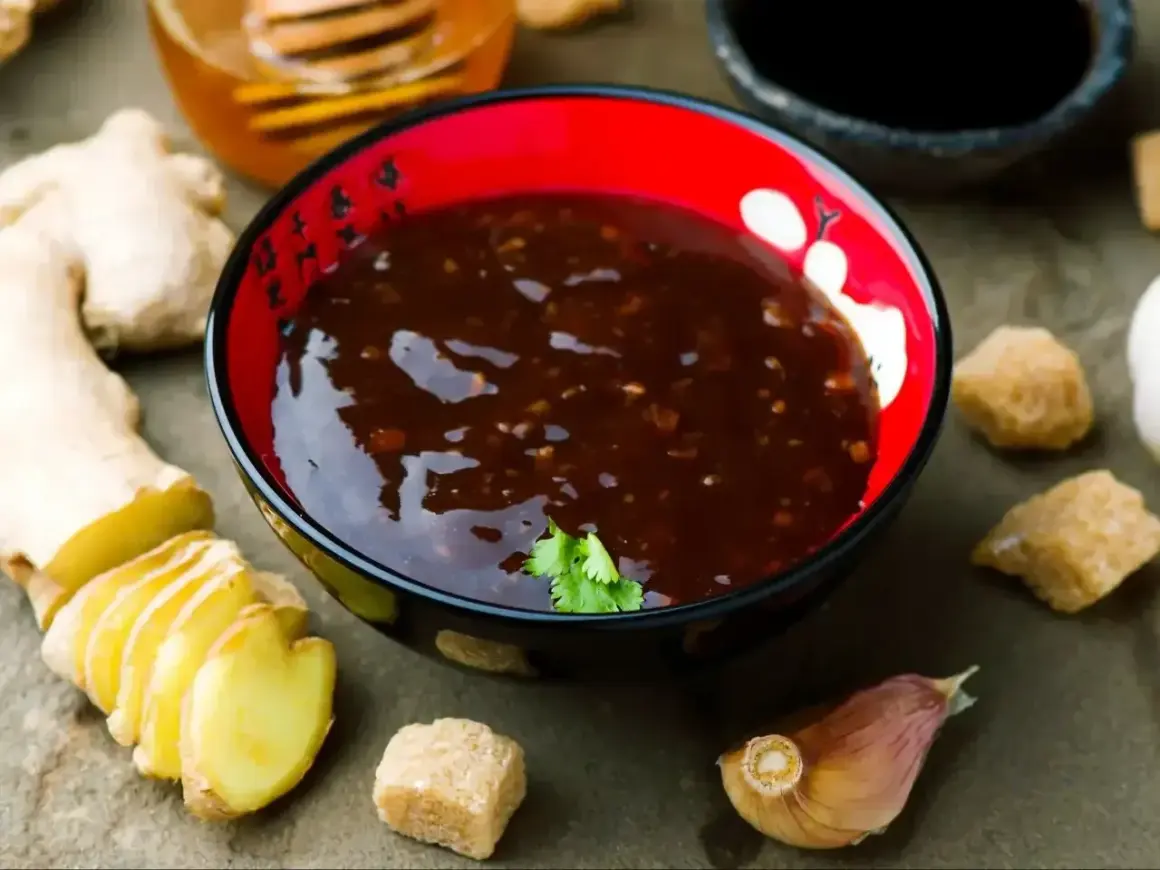 Sos teriyaki przepis: miseczka pełna aromatycznego sosu, obok świeży imbir, czosnek i brązowy cukier. Idealne składniki do domowego sosu teriyaki.