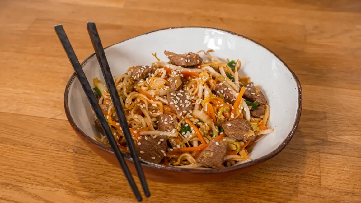 Makaron Chow Mein z wołowiną, warzywami i sezamem w misce z pałeczkami.