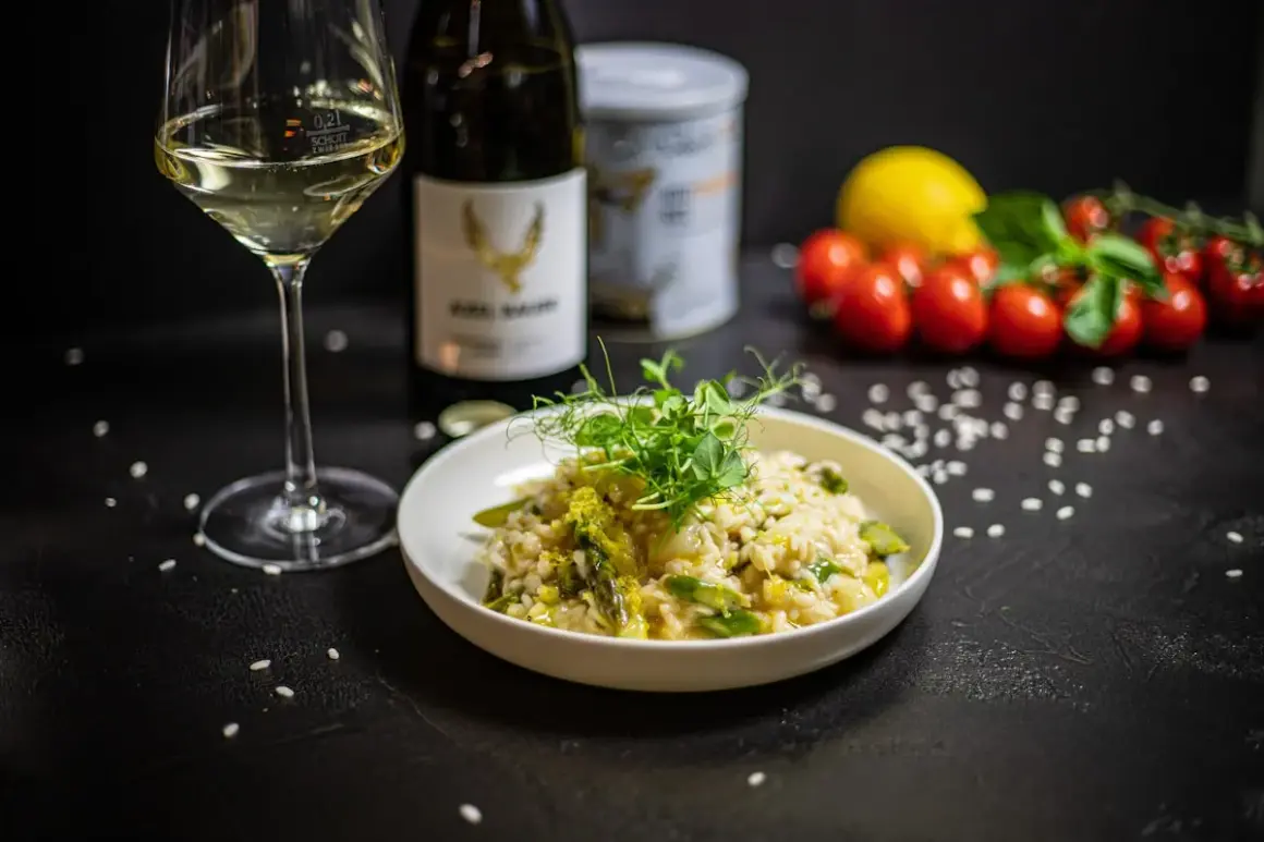 Klasyczne risotto z kurczakiem, podane z zielonymi szparagami i kiełkami, obok kieliszka białego wina i butelki.