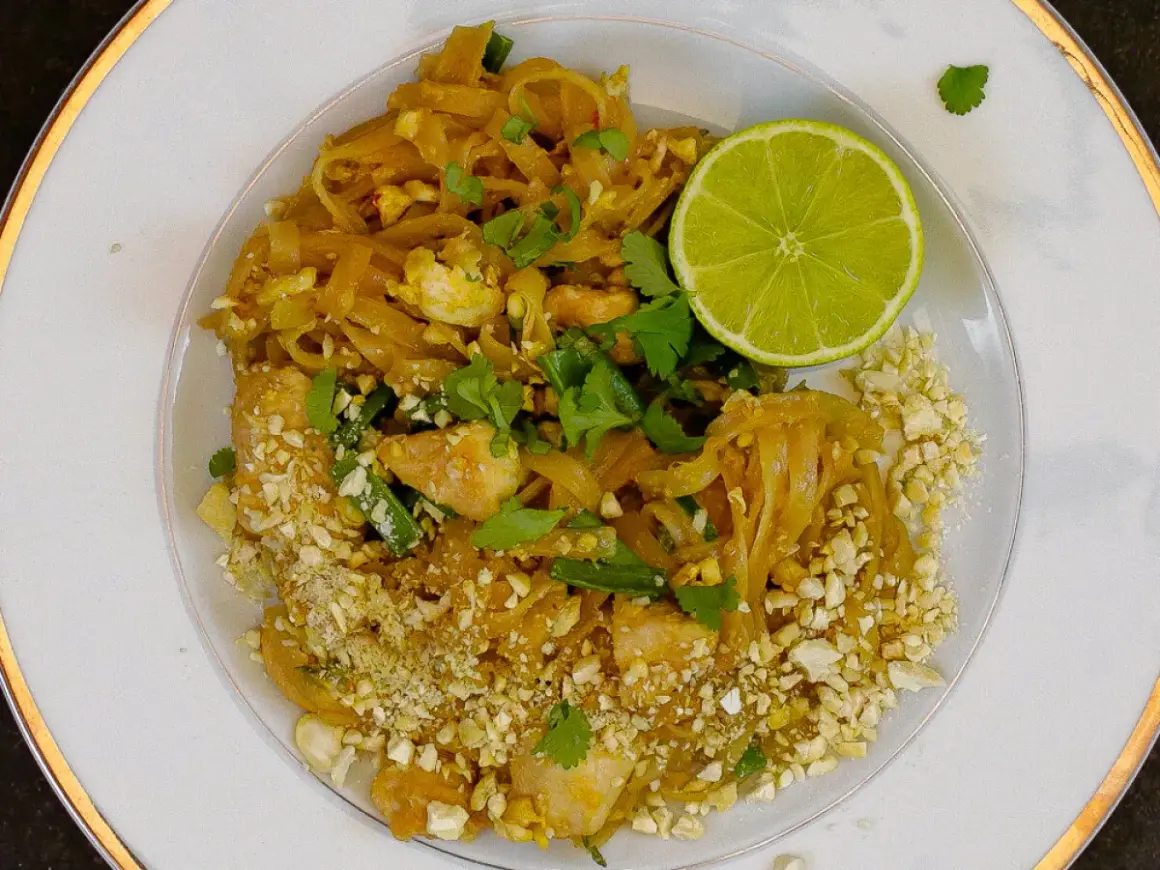 Talerz z Pad Thai, posypany orzeszkami ziemnymi, z połówką limonki i kolendrą.