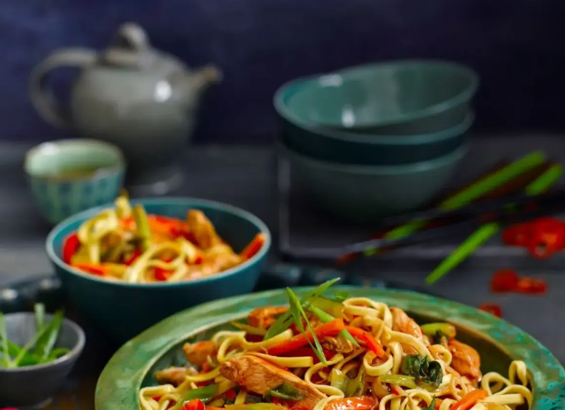 Makaron Chow Mein z kurczakiem i warzywami w misce, na tle czajniczka i pałeczek.
