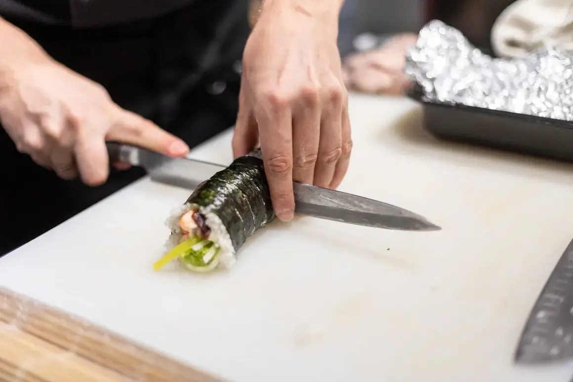 Dłonie kroją rolkę sushi. Zastanawiasz się, czy sushi można zamrozic? Ten obrazek pokazuje przygotowanie świeżego sushi.