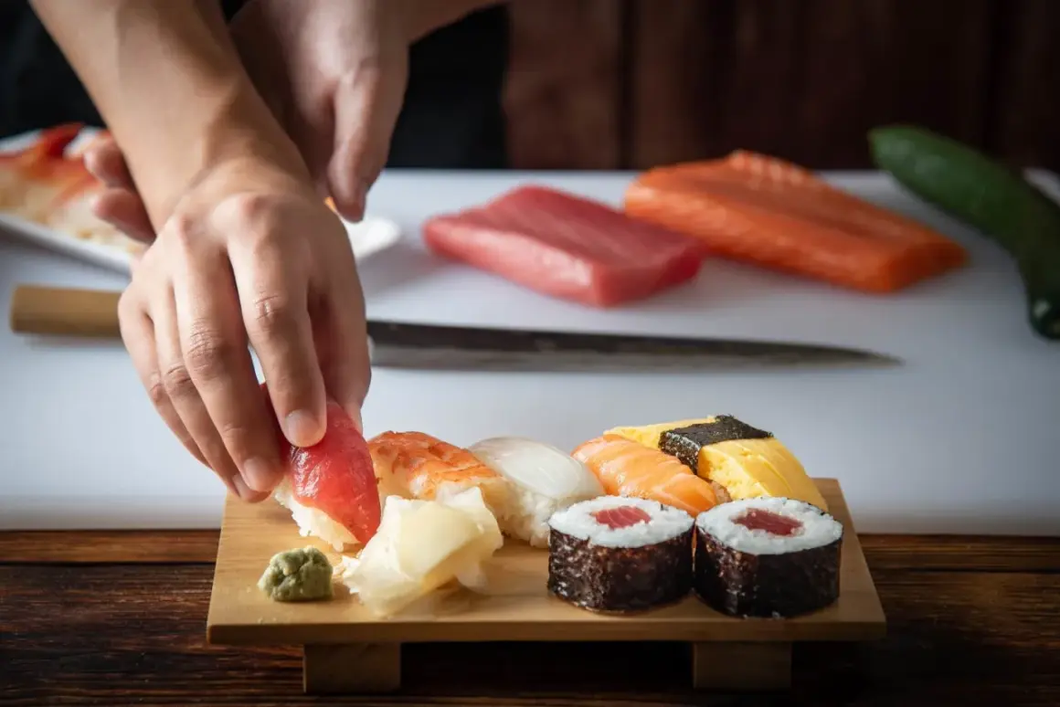 Ręce układają kawałek tuńczyka na sushi. Na desce widać też krewetkę, kalmara, łososia, omlet i rolki. Idealne składniki, by dodać do sushi.