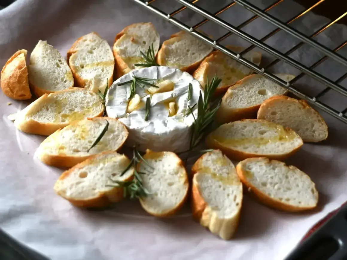 Pyszny, pieczony camembert z czosnkiem i rozmarynem, otoczony chrupiącymi grzankami skropionymi oliwą. Idealna przekąska.