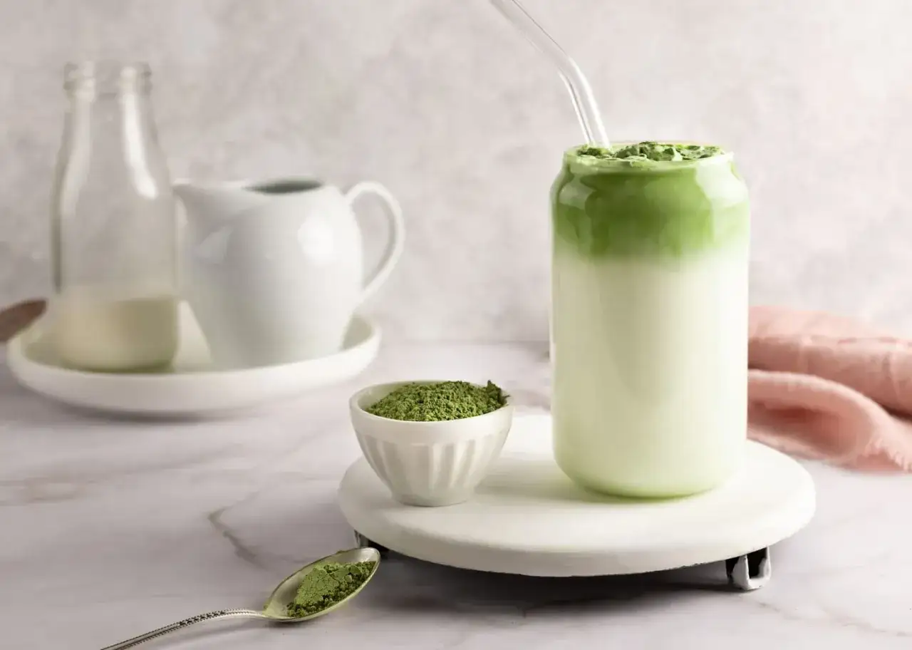 Jak zrobić matcha latte? Na stole mleko, proszek matcha w miseczce i łyżeczce, oraz gotowy napój w szklance ze słomką.