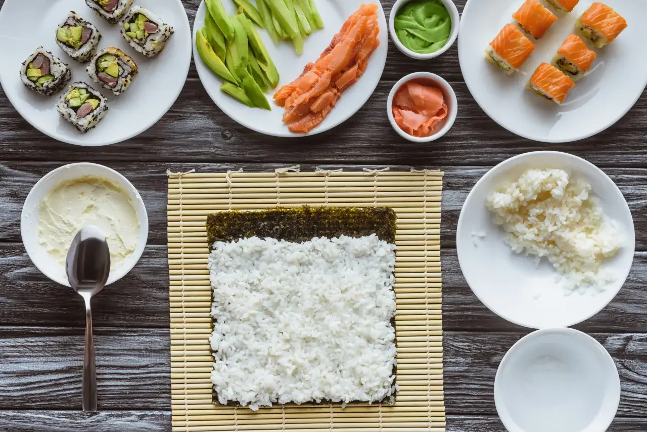 Jak ugotować ryż do sushi? Na macie bambusowej ułożono nori i ryż. Obok składniki: łosoś, awokado, wasabi, imbir, serek.