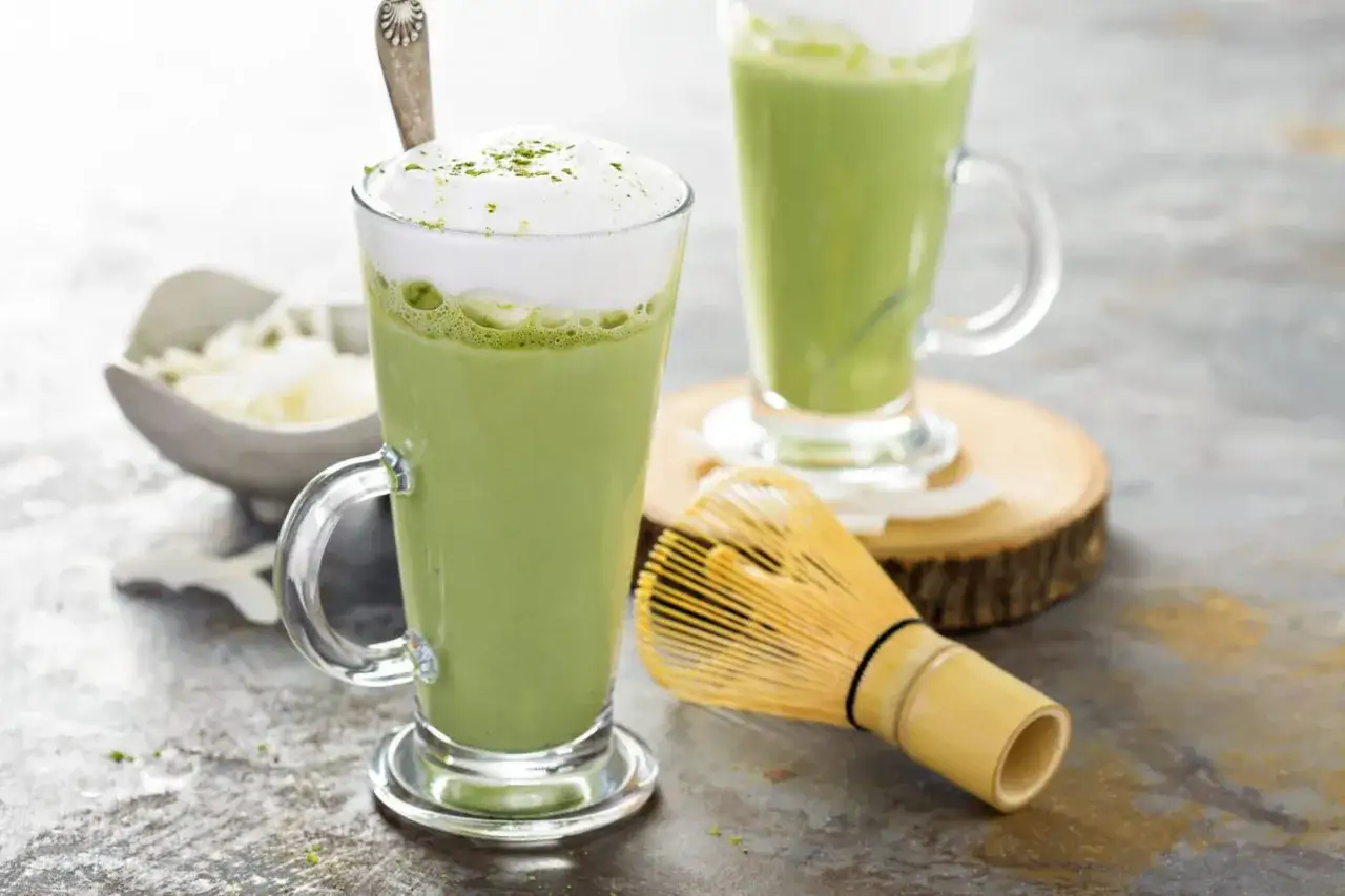 Zielony, spieniony napój w szklance, czyli matcha latte. Obok bambusowa miotełka do ubijania.