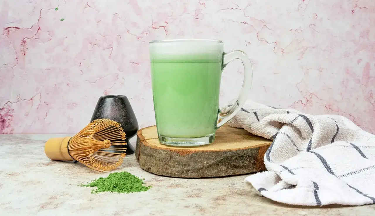 Zielony, spieniony matcha latte w szklance, obok bambusowa miotełka i proszek matcha. Idealny napój, jeśli zastanawiasz się, matcha latte gdzie kupić.