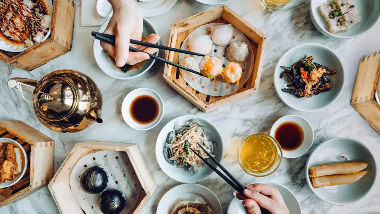 Stół pełen pysznego dim sum: pierożki, bułeczki, sajgonki i złoty dzbanek z herbatą.