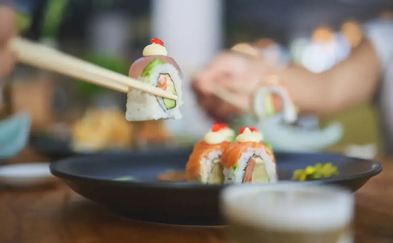 Chopsticks unoszą rolkę sushi z łososiem i awokado. Na talerzu czekają kolejne porcje, idealne do jedzenia z czym sushi.