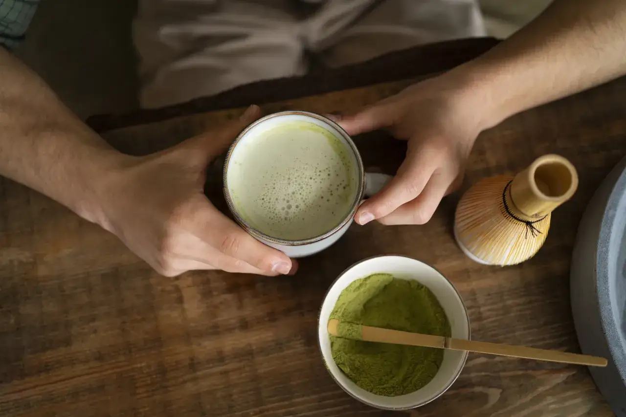 Dłonie trzymają kubek z zielonym napojem. Obok miseczka z proszkiem matcha i bambusowa miotełka. Czy matcha odchudza?