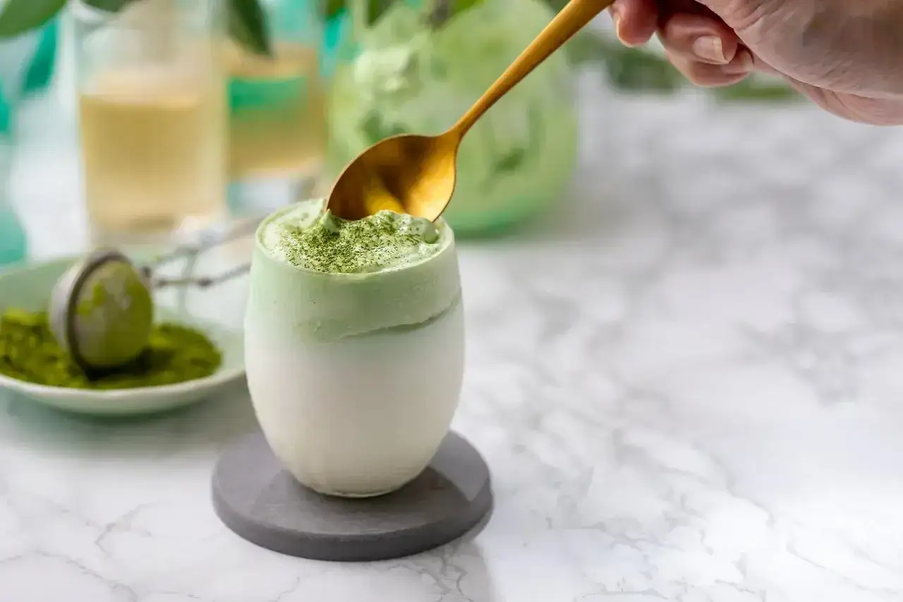 Puszysty matcha latte, czyli jak zrobić ten popularny napój. Złota łyżka zanurza się w zielonej piance.