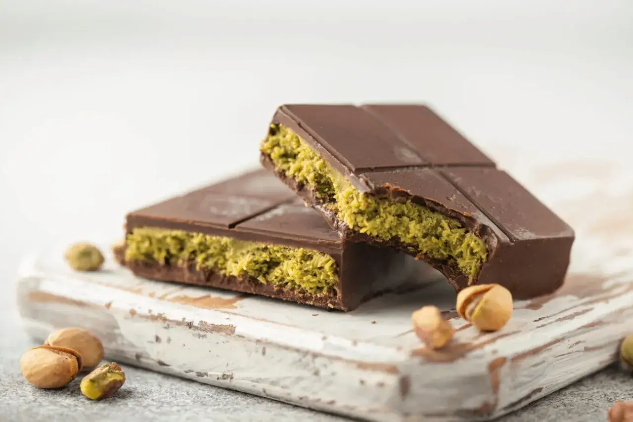Czekoladowe batony z nadzieniem z pistacji, przypominające ciasto kataifi. Obok leżą łuskane i niełuskane pistacje.